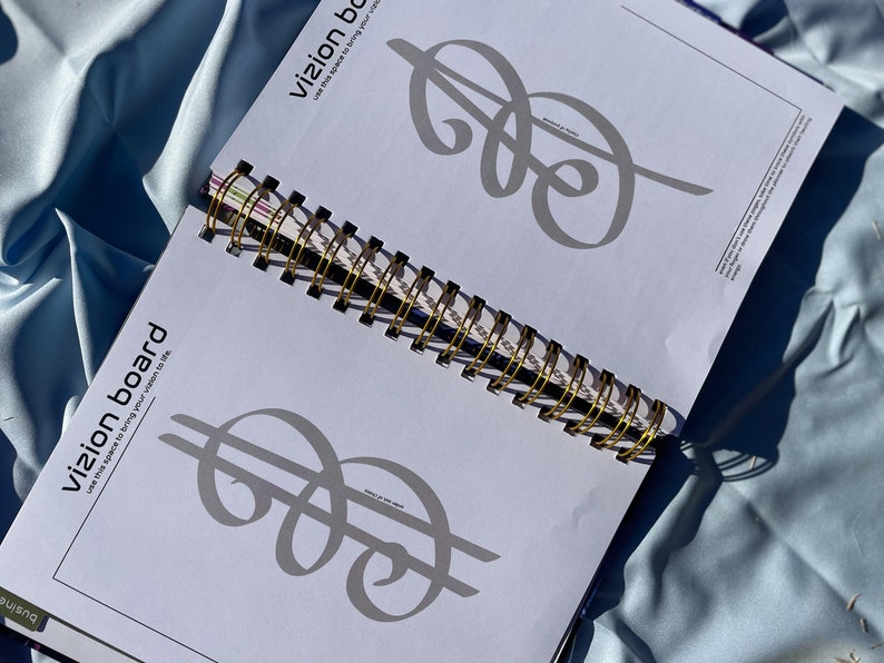 Pode incluir: Caderno espiral aberto com encaderna&ccedil;&atilde;o dourada. As p&aacute;ginas exibem um design de sinal de d&oacute;lar cinza e as palavras "vizion board" e "use this space to bring your vision to life". O caderno est&aacute; em tecido azul claro.