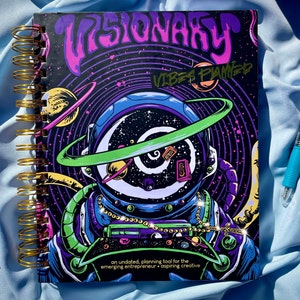 Pode incluir: Um planejador espiralado com uma capa vibrante com tema c&oacute;smico. O design apresenta um astronauta com um capacete cheio de gal&aacute;xias, planetas e o texto "VISIONARY VIBES PLANNED". &Eacute; uma ferramenta de planejamento sem data.