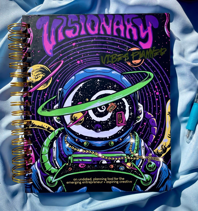 Pode incluir: Um planejador espiralado com uma capa vibrante com tema c&oacute;smico. O design apresenta um astronauta com um capacete cheio de gal&aacute;xias, planetas e o texto "VISIONARY VIBES PLANNED". &Eacute; uma ferramenta de planejamento sem data.