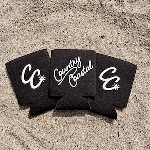 May include: Three black foam can coolers with white lettering. The middle cooler has the text "Country Coastal" written on it. The cooler on the left has the letter "C" with a star next to it. The cooler on the right has the letter "C" with a star next to it.