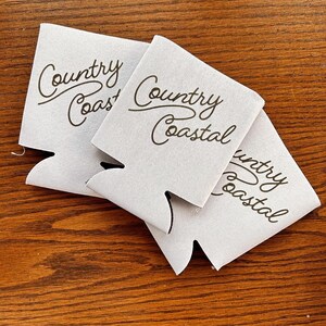 May include: Three white foam beverage insulators with the text "Country Coast" and "Country Coastal" printed in brown.