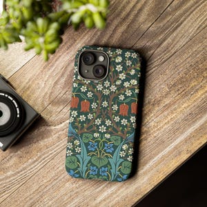 May include: A smartphone case with a floral pattern featuring white flowers, red fruit, and blue foliage on a dark green background. The case is on a wooden surface, with a camera and greenery in the background.