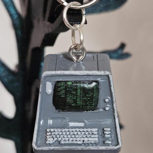 May include: A gray miniature computer keychain with a silver keyring. The computer has a screen displaying green text, a keyboard, and buttons. The keychain is a retro-style accessory.