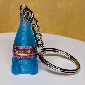 May include: A blue bottle-shaped keychain with a silver chain and keyring. The bottle has a label with the word "Quantum" on it. The keychain is made of a 3D printed material.