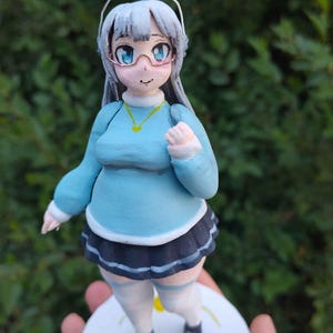 May include: A hand-painted clay figurine of an anime-style character with long silver hair, wearing a light blue long-sleeved sweater, a dark pleated skirt, and white thigh-high socks. The figure has glasses, a gold necklace, and stands on a round base.