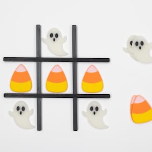 May include: A Halloween-themed tic-tac-toe game with candy corn and ghost pieces. The game board is black with white lines. The candy corn pieces are orange, yellow, and white. The ghost pieces are white with black eyes.