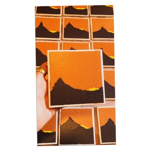 Könnte beinhalten: Eine Sammlung quadratischer Untersetzer in Orange und Schwarz mit einer Silhouette eines Berges und einem gelben Sonnenuntergang. Die Untersetzer sind gestapelt und in einem Gittermuster angeordnet.