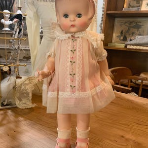 May include: A vintage doll with a pink dress, white lace collar, and matching shoes. The doll has a pink bow in its hair and is standing on a wooden surface. The dress features embroidered floral details.