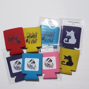 May include: A collection of koozie coolers in various colors, including red, yellow, blue, navy, and pink. Each cooler features a different cat or dog-themed graphic and text. The text includes phrases like "Crazy Cat Lady" and "A Dog Has An Owner, A Cat Has A Staff."