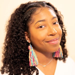 May include: A woman with dark curly hair smiles, wearing colorful tassel earrings. The earrings feature strands of pink, yellow, blue, and green. She wears a silver necklace with two small, round pendants. The background is a solid, light beige color.