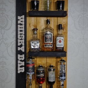 Może przedstawiać: Drewniany bar zamontowany na ścianie z napisem "Whiskey Bar" napisanym pionowo po lewej stronie. Bar ma dwie półki z różnymi butelkami alkoholu, w tym Jack Daniels, Label 5, Jim Beam, Captain Morgan, Republic, Gran Imperio i Borowa. Butelki są zamontowane na systemie dozowania z dyszami.
