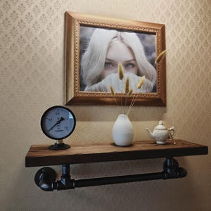 May include: A wooden shelf with black pipe supports, displaying a framed picture, a gauge, a white vase with dried plants, and a small teapot. The picture shows a woman with blonde hair. The shelf is mounted on a patterned wall.