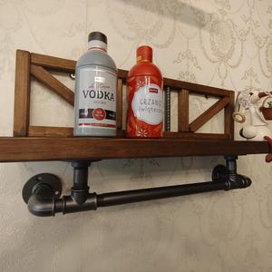 Puede incluir: Estante de madera oscura con dos botellas y una figura decorativa. El estante tiene un toallero de tuberías negras. Las botellas están etiquetadas como "Vodka" y "Grzanie świąteczne".
