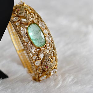 Könnte beinhalten: Ein goldfarbenes Armband mit einem ovalen, hellgrünen Edelstein in der Mitte. Das Armband ist mit kleinen, klaren Edelsteinen besetzt und weist verzierte, detaillierte Designs auf. Kleine rote Edelsteine sind ebenfalls sichtbar.