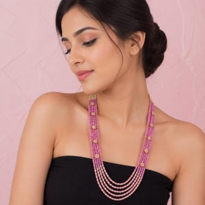 Op de afbeelding: Een meerstrengs ketting met vijf rijen kleine, gefacetteerde roze kralen. De strengen zijn verbonden door kleine, goudkleurige bloemvormige tussenstukjes met witte centra, wat zorgt voor een gelaagd ontwerp dat in een cascade op de borst rust.
