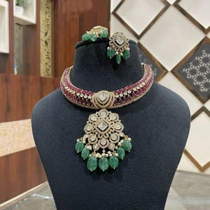Könnte beinhalten: Ein luxuriöses Schmuckset mit Halskette und Ohrringen. Die Halskette hat ein rot-goldenes Band mit einem großen Anhänger, der mit klaren und grünen Edelsteinen verziert ist. Die Ohrringe haben ein ähnliches Design mit grünen Edelstein-Tropfen.