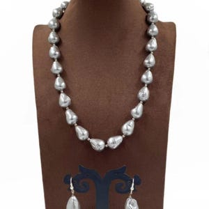 May include: A silver-colored pearl necklace and matching earrings set. The necklace features large, irregularly shaped pearls strung together. The earrings have a similar design, with a single pearl suspended from a hook. The jewelry is displayed on a brown jewelry bust.