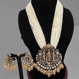 May include: An ornate necklace and earring set. The necklace features a multi-strand ivory bead design with a large, detailed pendant. The pendant has a gold-toned frame with figures and clear and peach-colored accents. Matching earrings complete the set.
