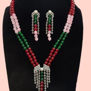 May include: A beaded necklace and earring set. The necklace features red, green, and pink beads. The earrings and necklace pendant have a silver-colored design with clear stones and a pearl accent. The jewelry is displayed on a black stand.