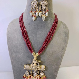 May include: A gold-toned necklace and matching earrings set. The necklace features a multi-strand burgundy beaded chain with a detailed pendant. The pendant has a figure seated on a decorative bar. The earrings have a similar design with white teardrop pearls.