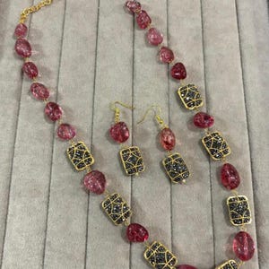 Könnte beinhalten: Ein Halsketten- und Ohrring-Set mit rosa und schwarzen Edelsteinperlen. Die Halskette hat eine goldene Kette und ist mit rosa, ovalen Perlen und rechteckigen, goldgerahmten schwarzen Edelsteinperlen verziert. Die Ohrringe spiegeln das Design der Halskette wider.