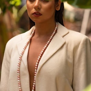 Puede incluir: Un largo collar de perlas rosas y blancas sobre una blazer color crema. Las perlas son una mezcla de rosa y blanco, creando un look suave y elegante. La blazer tiene un corte clásico y un color neutro.
