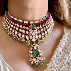 Könnte beinhalten: Eine luxuriöse Halskette mit einem tief burgunderfarbenen Band, verziert mit weißen Edelsteinen und goldenen Akzenten. Die Halskette hat einen großen smaragdgrünen Anhänger und kleinere grüne Edelstein-Tropfen, die ein elegantes Design schaffen.