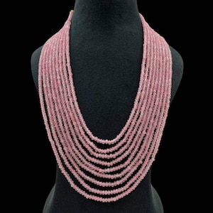 May include: A multi-strand necklace displayed on a black mannequin. The necklace features numerous strands of small, round, light pink beads. The beads are evenly spaced, creating a cascading effect. The background is a solid black.