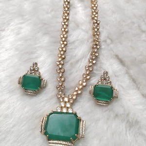 Könnte beinhalten: Ein goldfarbenes Schmuckset mit einer Halskette und passenden Ohrringen. Die Halskette hat einen großen, smaragdgrünen Anhänger und eine zarte Kette. Die Ohrringe und der Anhänger haben einen rechteckigen smaragdgrünen Stein, der von kleinen, klaren Steinen umgeben ist.