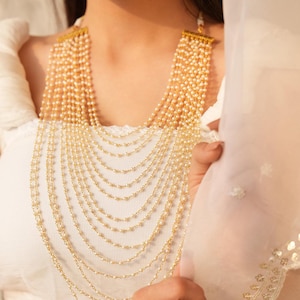 May include: A multi-strand pearl necklace with gold-toned accents. The necklace features numerous strands of small, round pearls cascading down the chest. The necklace is worn over a white top, creating a contrast with the pearls' warm tones.