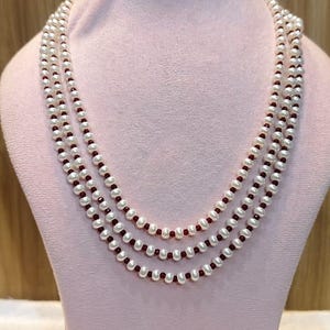 Puede incluir: Collar de perlas de tres hilos con perlas blancas y pequeñas cuentas rojas alternas. El collar se exhibe sobre un busto de maniquí rosa sobre un fondo marrón claro.