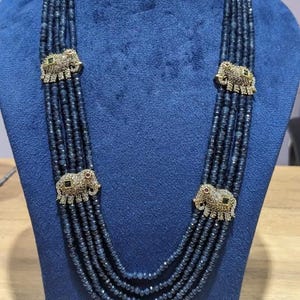 May include: A multi-strand necklace with dark blue beads and gold-colored elephant charms. The necklace is displayed on a blue bust. Each elephant charm has small, colorful accents.