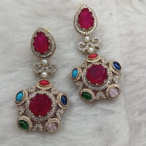 Könnte beinhalten: Ein Paar goldfarbene, sternförmige Ohrringe mit einem großen, runden, rubinroten Mittelstein. Jeder Ohrring hat kleinere, farbige Edelsteine in Türkis, Grün, Blau und Rosa. Die Ohrringe haben einen tropfenförmigen, rubinroten Stein oben, mit einem Perlenakzent.