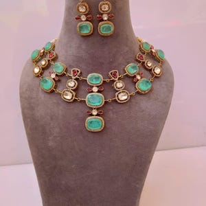 Könnte beinhalten: Ein goldfarbenes Halsketten- und Ohrring-Set mit smaragdgrünen und rubinroten Edelsteinen. Die Halskette hat mehrere Reihen ovaler grüner Steine und kleinere rote Akzente. Die Ohrringe spiegeln das Design der Halskette wider.