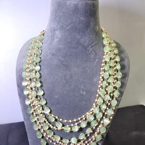 Op de afbeelding: Een meerrijige ketting tentoongesteld op een grijze mannequin. De ketting heeft strengen lichtgroene ovale kralen en kleine witte kralen, verbonden door goudkleurige kettingen. De ketting is een statement piece.