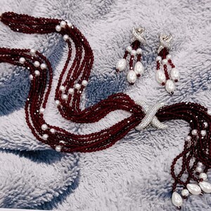 May include: A burgundy beaded necklace and matching earrings set. The necklace features multiple strands of small, dark red beads interspersed with white pearls. The earrings have a similar design, with pearl drops and silver accents. The jewelry is displayed on a textured, light gray surface.