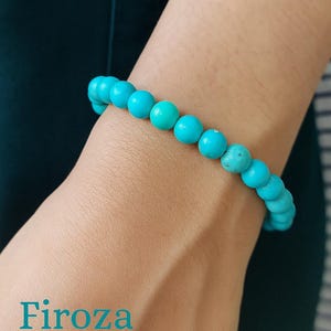 Pode incluir: Uma pulseira de contas turquesa é usada no pulso. A pulseira é feita de contas redondas e polidas em uma vibrante cor azul-esverdeada. A palavra "Firoza" está impressa em turquesa na pele.