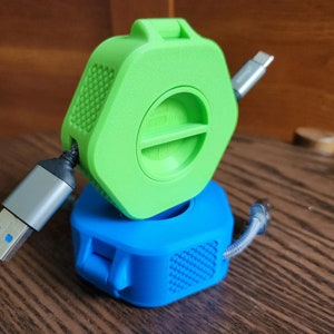 Puede incluir: Un organizador de cables hexagonal de color verde y azul. El organizador tiene una ranura para enrollar un cable y una pequeña abertura para que el cable pase.