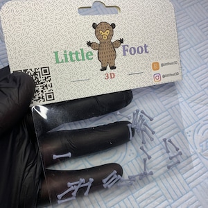 Può includere: Un pacchetto di piccole ossa di plastica bianca. La confezione è etichettata "Little Foot 3D" e presenta un orso dei cartoni animati. La confezione include anche le maniglie dei social media per l'azienda.