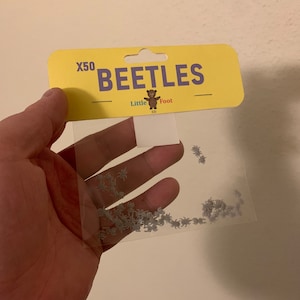 May include: A clear plastic bag containing 50 small grey plastic beetles. The bag has a yellow header with the text "X50 BEETLES" and a small brown bear logo with the text "Little Foot 3D".