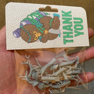 May include: A small plastic bag containing a collection of grey, unpainted, miniature tree branches. The bag is labelled with a thank you card featuring a cartoon bear wearing a green scarf.