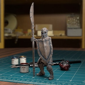以下が含まれることがあります： 槍と盾を持ったブロンズ色のミニチュア戦士のフィギュア。フィギュアは緑色のカッティングマットの上に立っており、20面ダイスが置かれている。