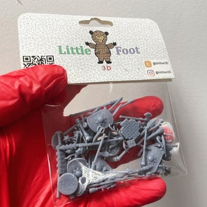 Può includere: Un sacchetto di plastica trasparente pieno di piccole armi e scudi in plastica grigia. Il sacchetto è etichettato "Little Foot 3D" con un logo di un orso dei cartoni animati e maniglie di social media.