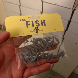 May include: A bag of 65 small, gray plastic fish. The bag has a yellow label with the text "X 65 FISH" and a logo for "Little Foot 3D".