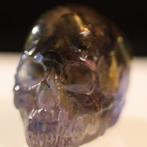 Puede incluir: Una escultura de cráneo translúcida e iridiscente con un tono marrón oscuro y morado. El cráneo está detallado con una cuenca del ojo visible y dientes.