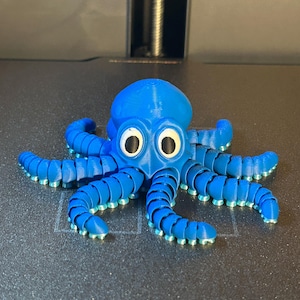 May include: A 3D-printed blue octopus toy with large, round eyes and segmented tentacles. The octopus has a playful design with a light blue gradient on the tentacle tips. The toy is sitting on a gray surface.