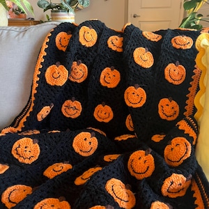 May include: A black crocheted blanket featuring numerous orange pumpkin faces with smiling expressions. The blanket has an orange border and is draped over a gray couch. A cozy, handmade Halloween-themed throw.