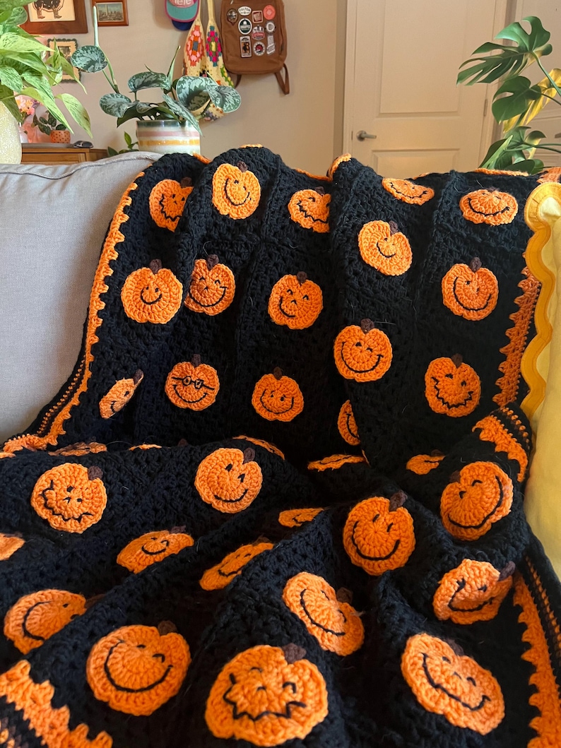 May include: A black crocheted blanket featuring numerous orange pumpkin faces with smiling expressions. The blanket has an orange border and is draped over a gray couch. A cozy, handmade Halloween-themed throw.