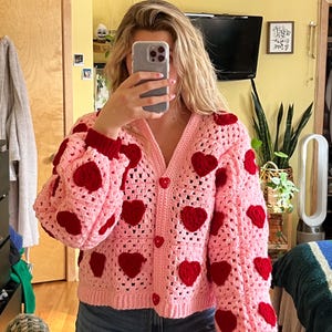 Puede incluir: Una chaqueta de punto de color rosa hecha a crochet con detalles de corazones rojos. La chaqueta tiene cuello en V y cierre de botones.