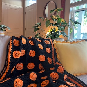 May include: A black and orange crocheted blanket featuring smiling pumpkin faces. The blanket has an orange border and is draped over a sofa. A yellow pillow with a scalloped edge is also on the sofa. The blanket is a festive Halloween decoration.